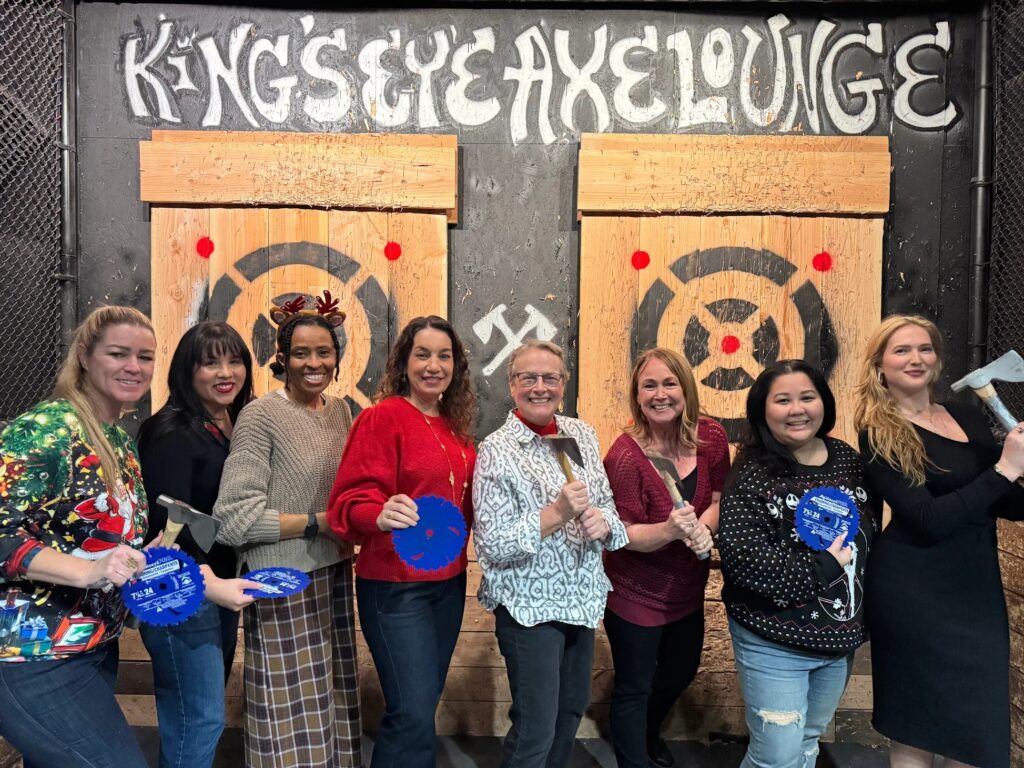 A group of eight women smiling and posing with axes and blue saw blades in front of wooden targets at King's Eye Axe Lounge in Long Beach, CA.